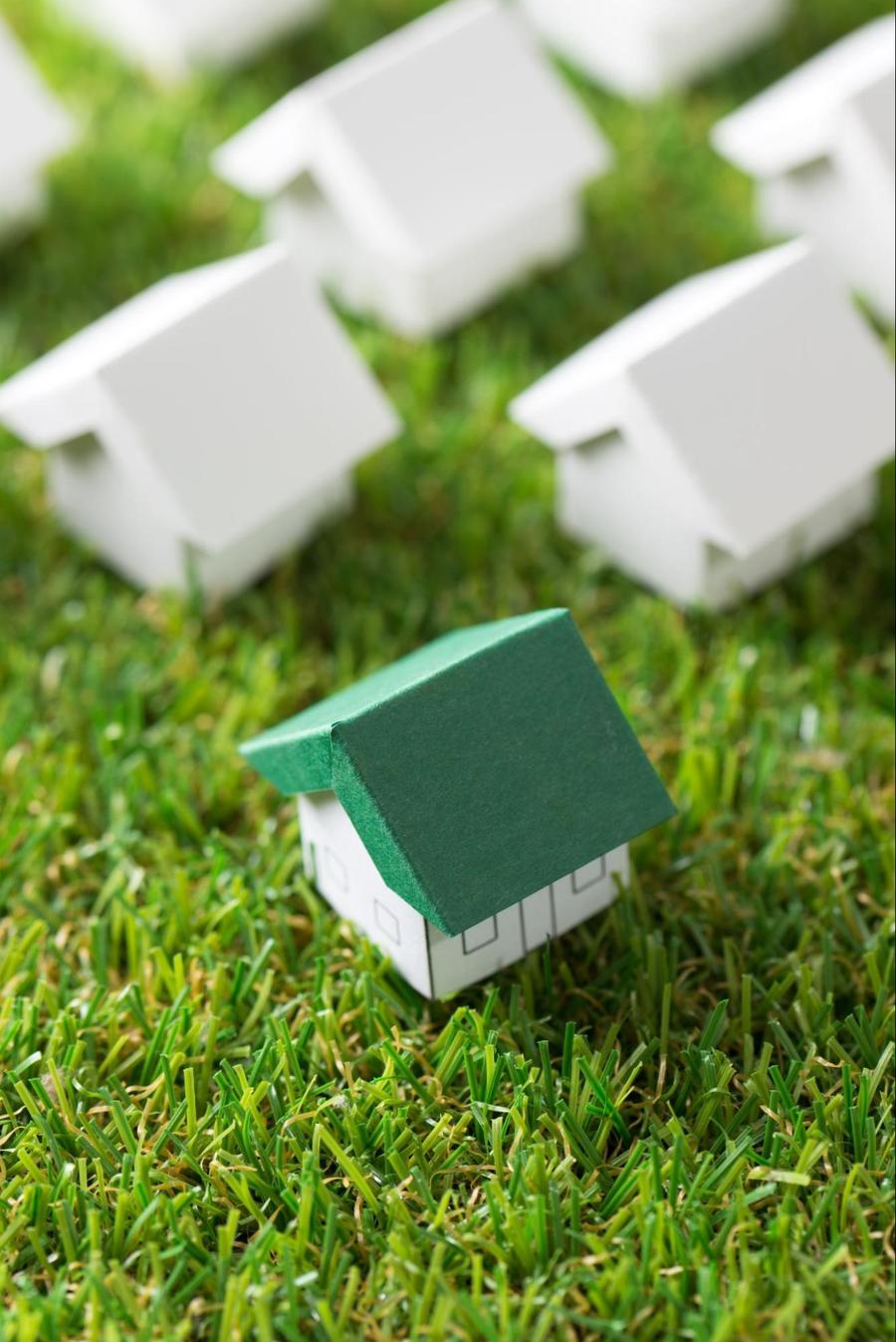 Close up of a miniature paper house with a green roof, sitting atop fake grass, and surrounded by 3 other mini white paper houses.