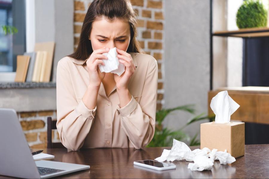 Brunette caucasian woman wearing a cream button blouse, sitting at a desk, blowing her nose