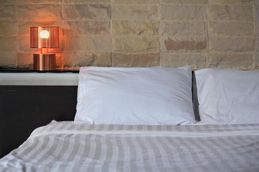 Bed with white and striped sheets with a stone wall behind it and a lamp on the nightstand