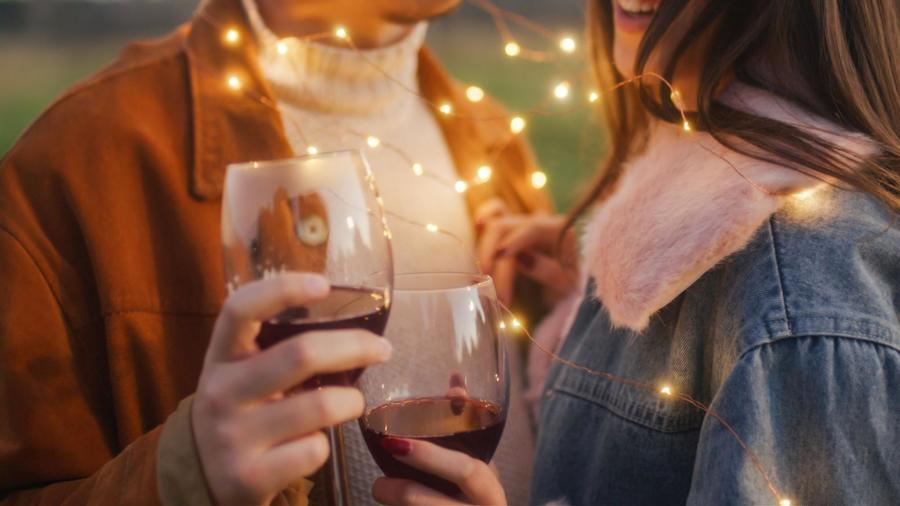 A couple holding wine glasses full of red wine, with LED fairy string lights wrapped around them.