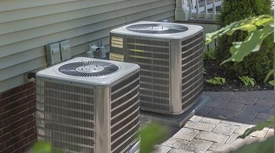 Two ground heat pump units sitting next to each other to the side of a house, atop a paved area, surrounded by greenery.