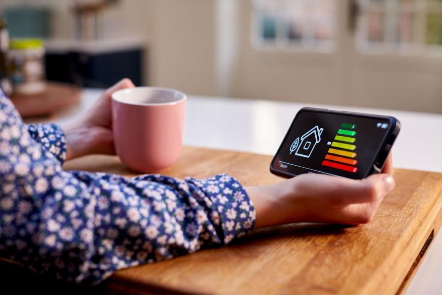 A person wearing a floral long sleeve shirt, holding a coral colored coffee mug in their left hand and their phone with a smart house app in the right hand.