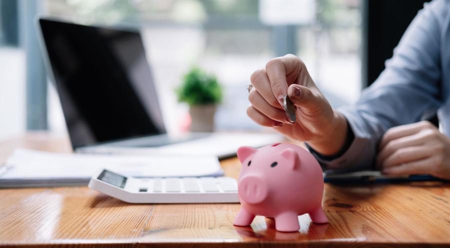 A close up of a small pink piggy bank sitting atop a wooden desk, next to a white calculator, stack of papers, and laptop with a person putting a coin in its slot.