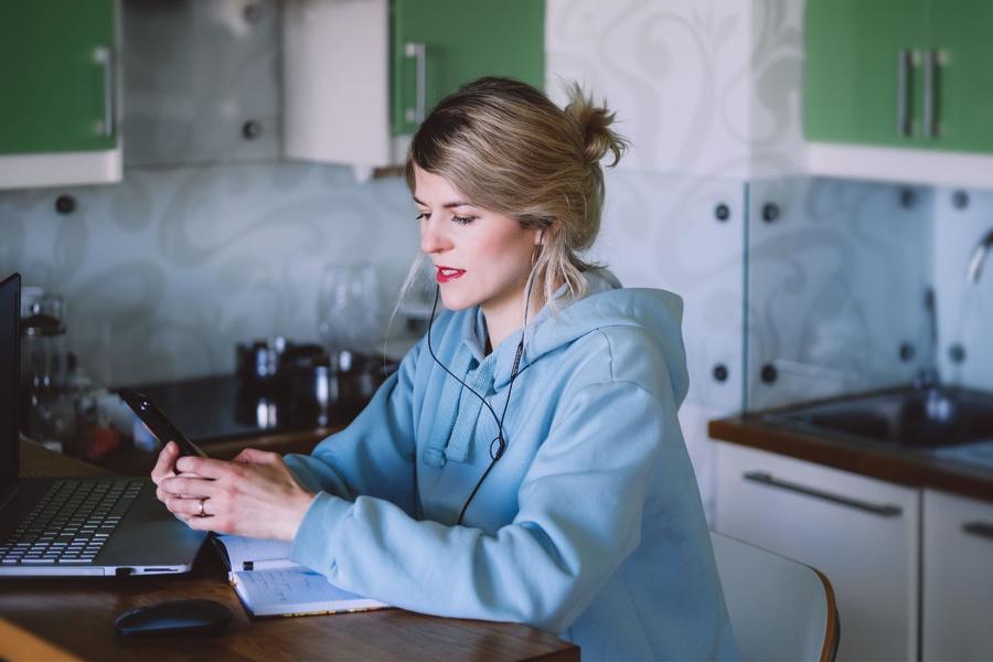 Woman with dirty blonde hair clipped up, wearing a sky blue hoodie, with black corded earbuds in, sitting at a kitchen table, looking at her phone in front of an open laptop and open lined notebook.