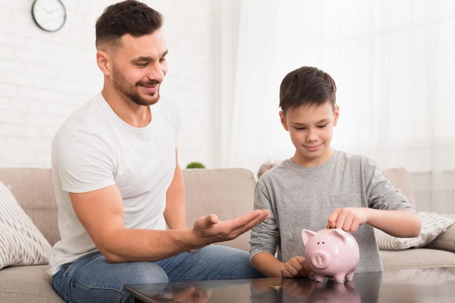A father and son sitting next to each other on the couch, with the father holding an open hand out while his son places coin in a pink piggy bank sitting on a coffee table in front of them.