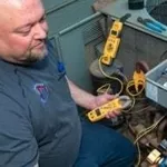 HVAC Tech wearing a Four Seasons branded shirt, working on an ac unit using multiple reading tools that are connected to the unit.