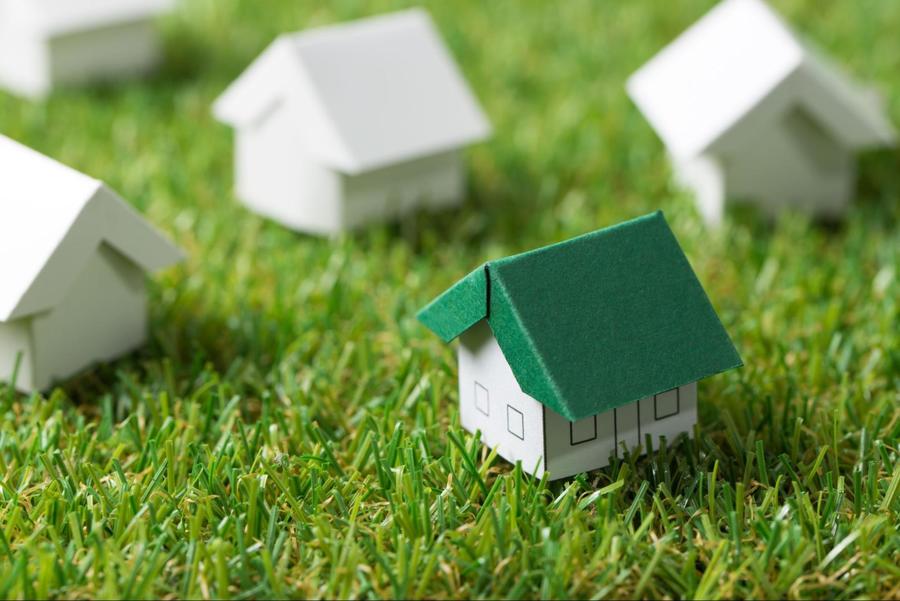 Close up of a miniature paper house with a green roof, sitting atop fake grass, and surrounded by 3 other mini white paper houses.