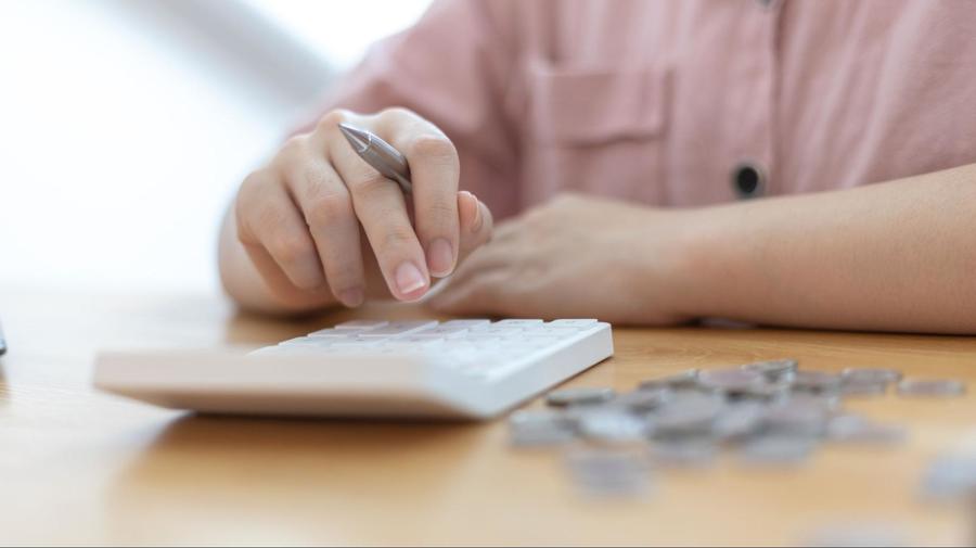 Someone holding a silver pen in their right hand while using a white calculator that is sitting on a wooden desk next to a pile of coins.