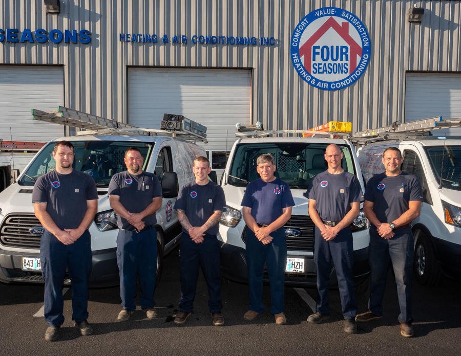 Four Seasons HVAC technicians in branded t-shirts standing in front of service vehicles parked in front of their warehouse